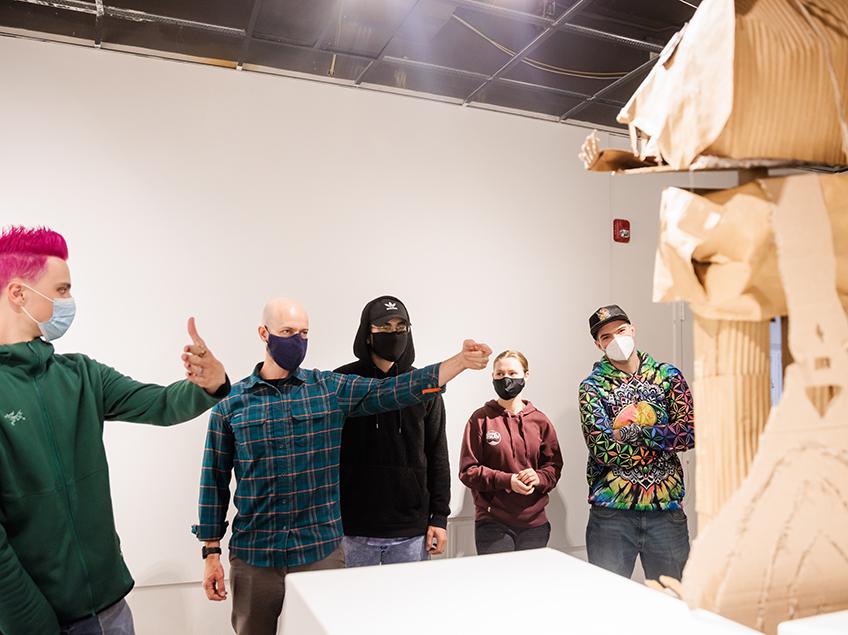 Professor Michael Bernhardt walks through a critique with his class in Gallery 199. By their hand gestures, they are discussing certain angles of the cardboard sculpture on a pedestal in the foreground. One student has electric pink hair.