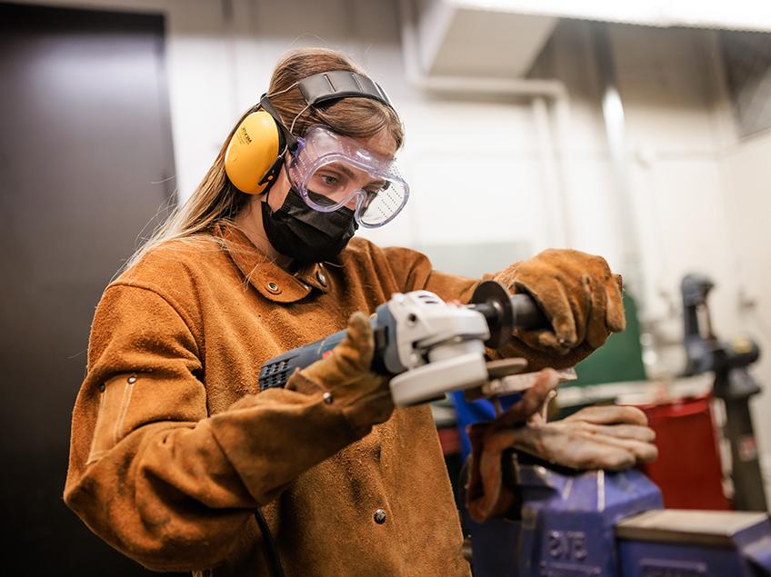 A student works with heavy machinery while wearing a heavy leather protective smock and gloves, along with soundproof headphones and safety goggles.