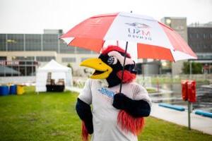 Rowdy, the MSU Denver University mascot, dressed as a roadrunner, stands on a large rock with arms raised and thumbs up. The scene is set on a grassy campus area with the Tivoli building and the Denver city skyline in the background.