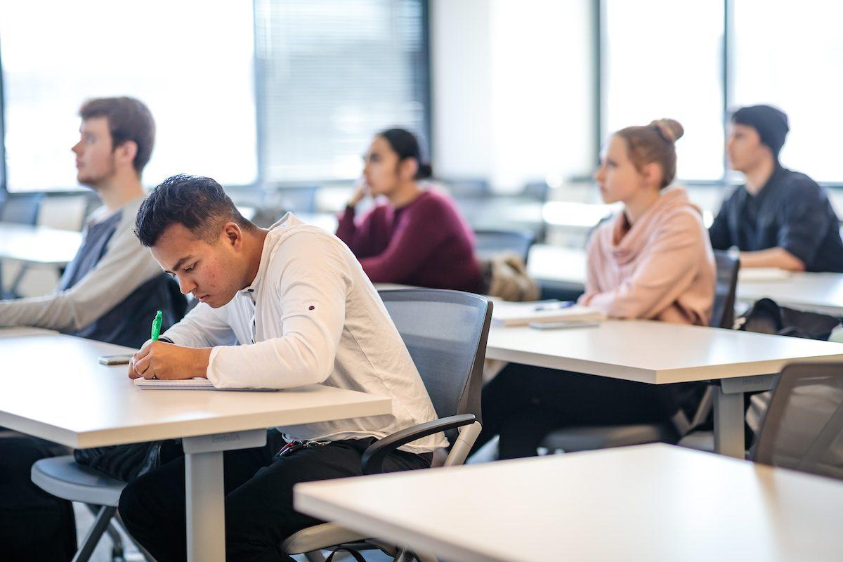 Several students sitting at desks in a classroom. The focus is on a student in the foreground wearing a white shirt and writing with a green pen. Other students in the background are attentively listening and taking notes.