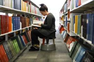 A college students sits on a stool in a library row reading a book.