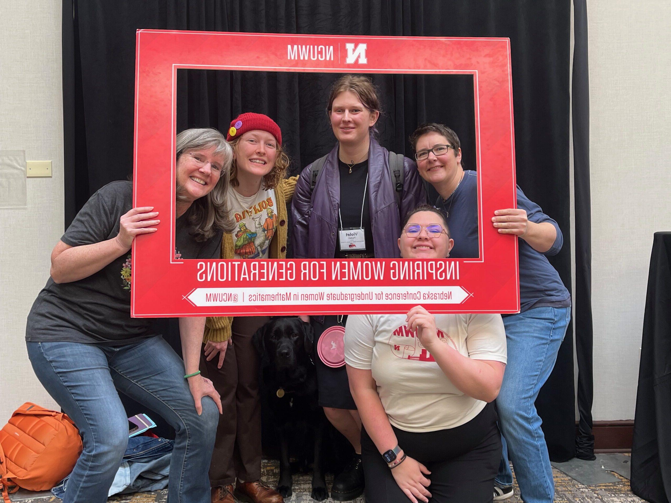 Three MSU Denver math students and Doctors Davis and McKenna all pose together in a giant picture frame at the 2025 Nebraska Conference for Undergraduate Women in Mathematics.